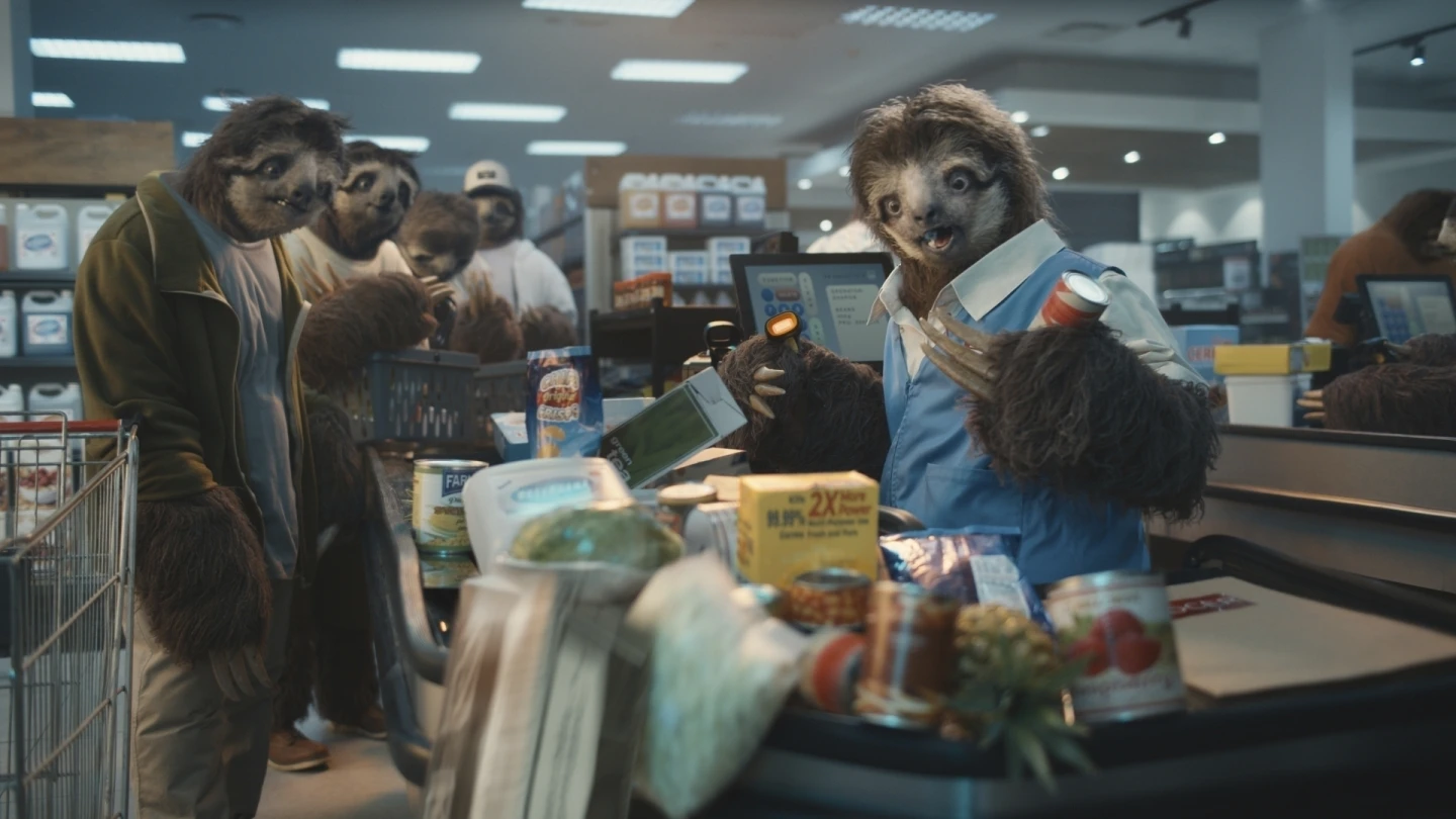 A humorous supermarket scene showing anthropomorphic sloths. A sloth dressed as a cashier scans groceries, holding a can in one hand and a barcode scanner in the other. Other sloths wait in line with their carts, surrounded by a variety of products, creating a lighthearted and quirky atmosphere.