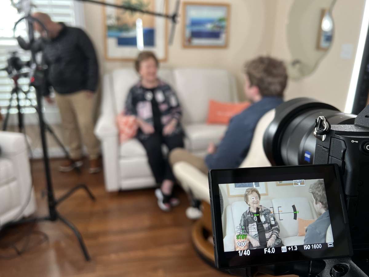 Behind-the-scenes shot of a video production setup, showing a woman being interviewed on a couch with a camera capturing her in focus. A crew member and equipment are visible in the background.